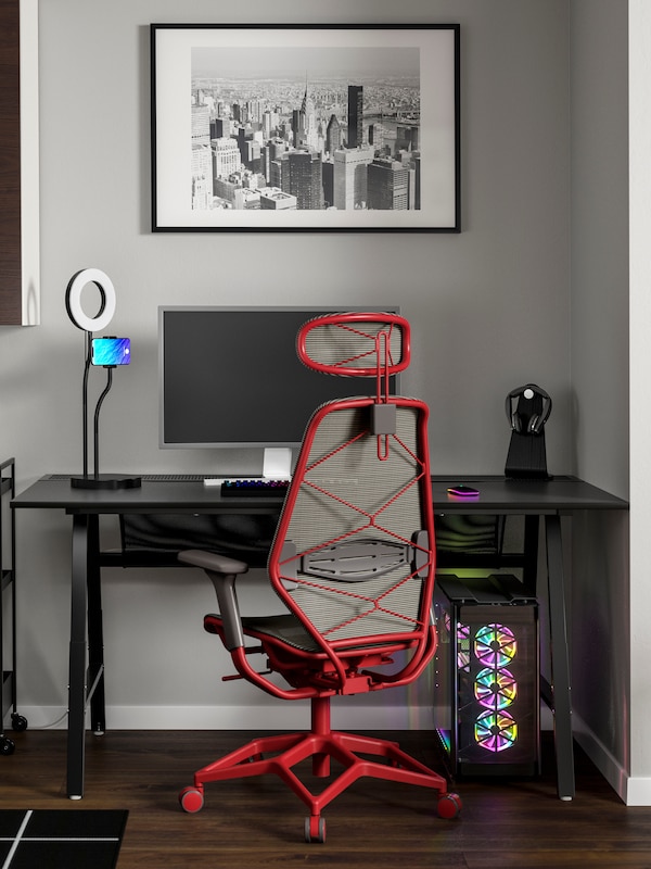An IKEA STYRSPEL gaming chair in grey and red in front of a black desk, in a room with grey walls and black and white artwork on the wall.