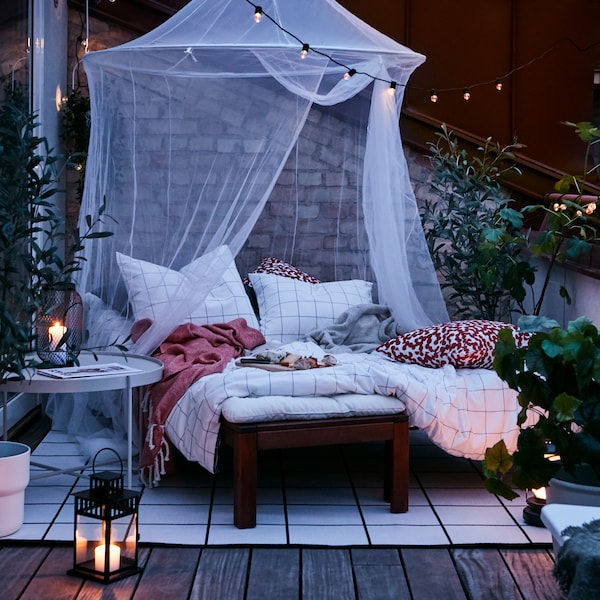 An outdoor terrace at dusk with a cosy nest made up of a lounging bed, SOLIG net, rug, lanterns and outdoor lighting chains.