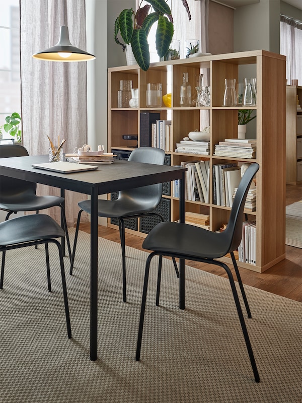 Black LIDÅS chairs and a black table sit on a rattan rug. The KALLAX shelving unit behind is being used as a room divider.