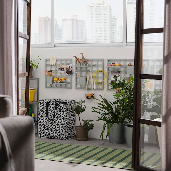 Doors open into a balcony with grey-green SNICKRA storage boards on a wall, a black/white bag, large potted plants and a rug.