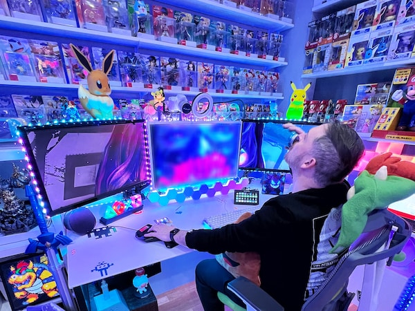 A man sitting on a desk playing video games on a computer with three screens, drinking in a can, in a room decorated with various figurines and lit with neon lights.