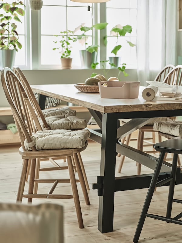 In a dining room are a large SKOGSTA wooden dining table with four chairs and several green plants in the window behind it.