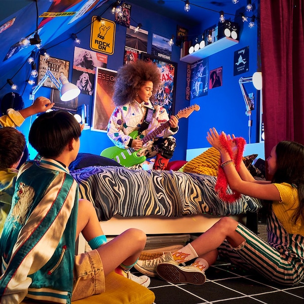 Four children in a blue room, three of them sitting on the floor watching the fourth child on a bed playing guitar like a rockstar. 