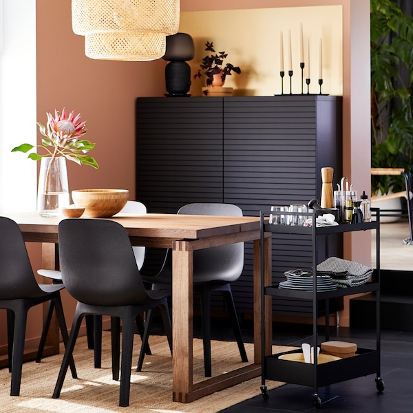 MÖRBYLÅNGA oak veneer table with black ODGER chairs by a window, with a black cabinet and a trolley holding tableware.