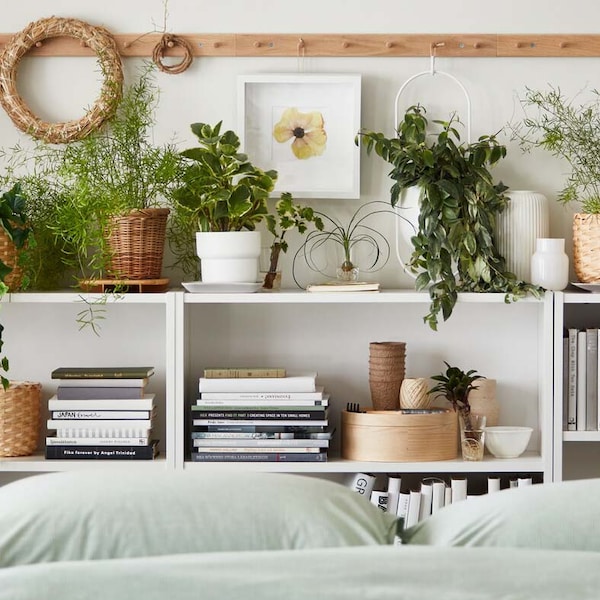 Multiple potted plants, real and artificial, stand amid books and decoration on shelves by a bed with BERGPALM bed linen.