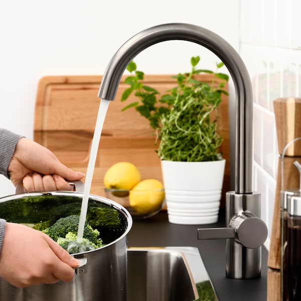 A person using an IKEA DELSJÖN kitchen mixer tap in dark grey stainless steel to rinse out vegetables in a pot.