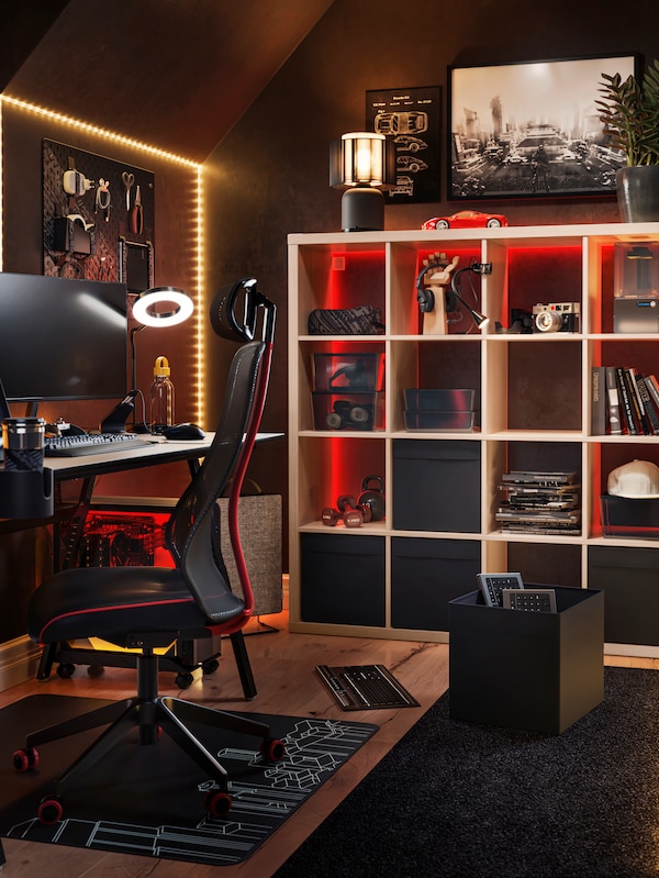 A dim gaming room with a desk and gaming chair, lit with red led strips, an IKEA KALLAX shelving unit in white in the back filled with various storage boxes and accessories.