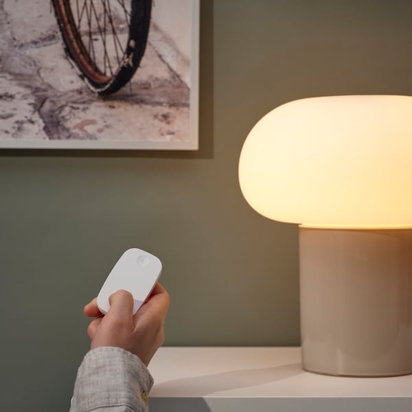 A hand holding a RODRET wireless dimmer/power switch at a lit lamp placed on a white table and next to a framed poster.