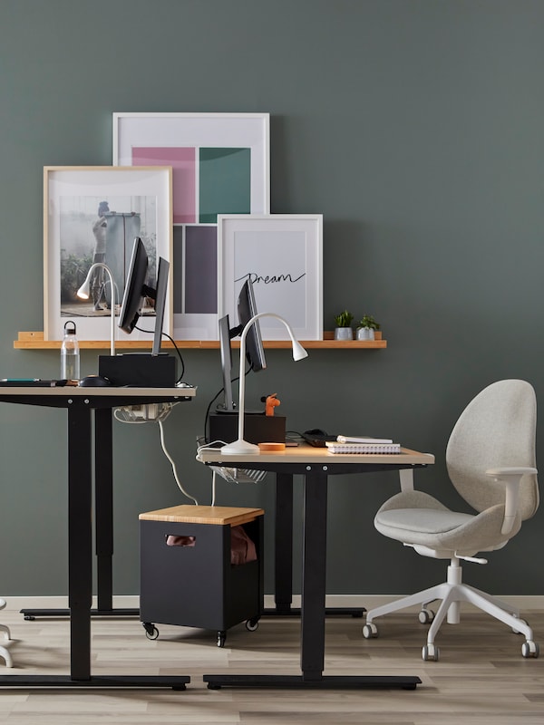 Two IKEA TROTTEN sit-stand desks in oak and black, a beige desk chair and an oak shelf on a green wall.