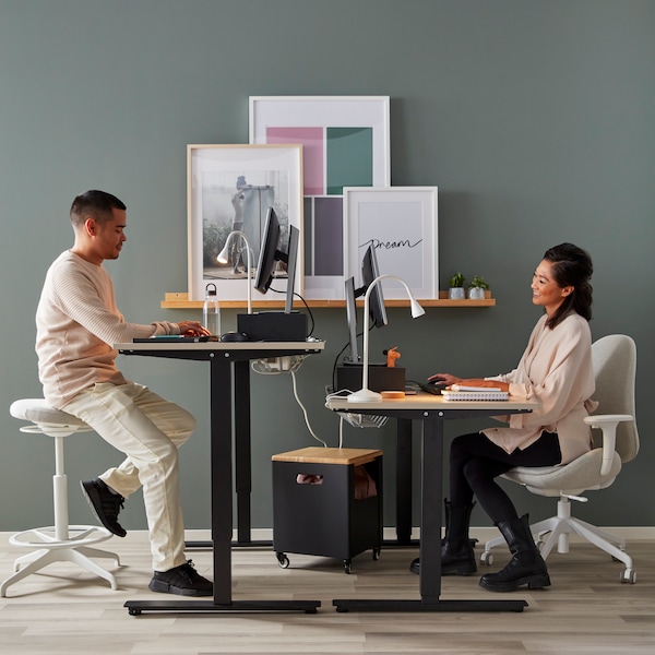 Two people sitting face to face on IKEA TROTTEN office desks in black, a green wall behind with picture frames on a picture ledge.