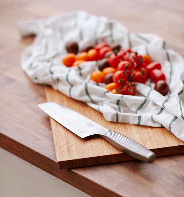 A vegetable knife in stainless steel on a wooden chopping board next to tomatoes on top of a tea towel.
