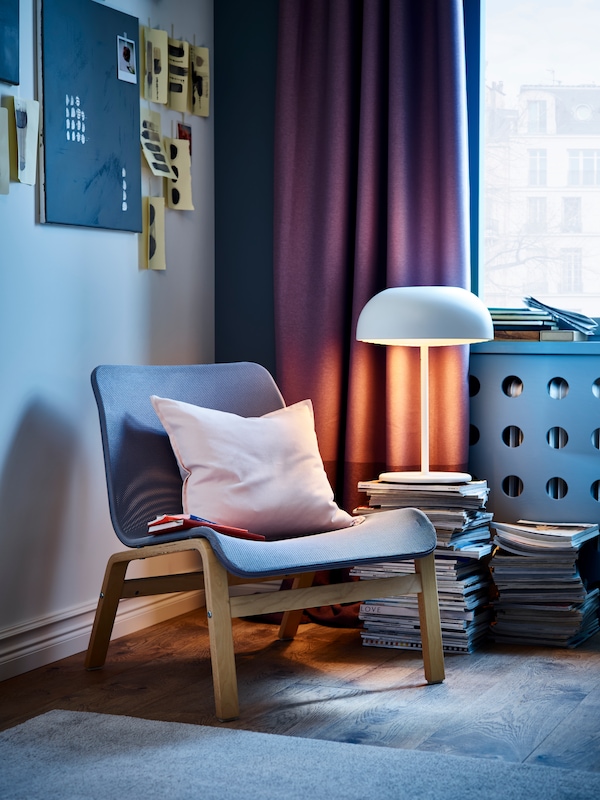 An IKEA NOLMYRA armchair in birch veneer and grey on a grey rug, a pink cushion over it and dark purple curtains on the window in the back.