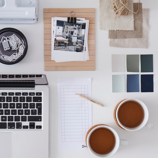 A white desk with a laptop, two cups of tea, a checklist, a wooden IKEA pencil and colour swatches.