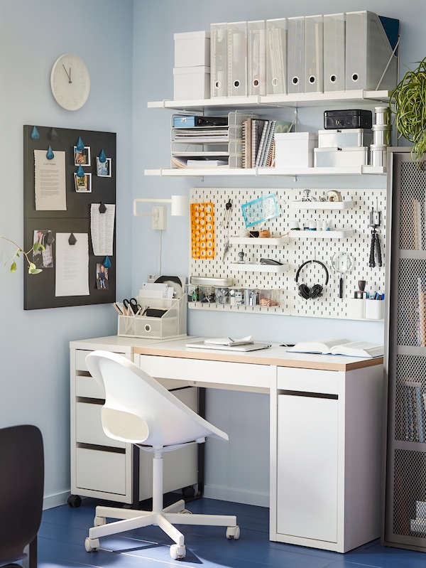 An IKEA MICKE white desk with a white desk chair, white shelves and pegboard on the wall above, filled with various office accessories and supplies.