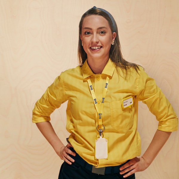 A woman with brown hair smiles while wearing a yellow IKEA co-worker collared shirt, lanyard and name tag.