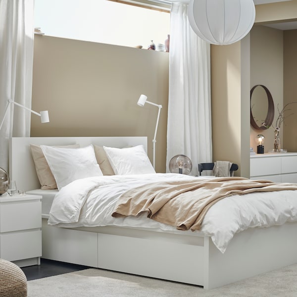 A high MALM bed with 4 storage boxes with a NYMÅNE floor/reading lamp on either side against a wall in a bright bedroom.