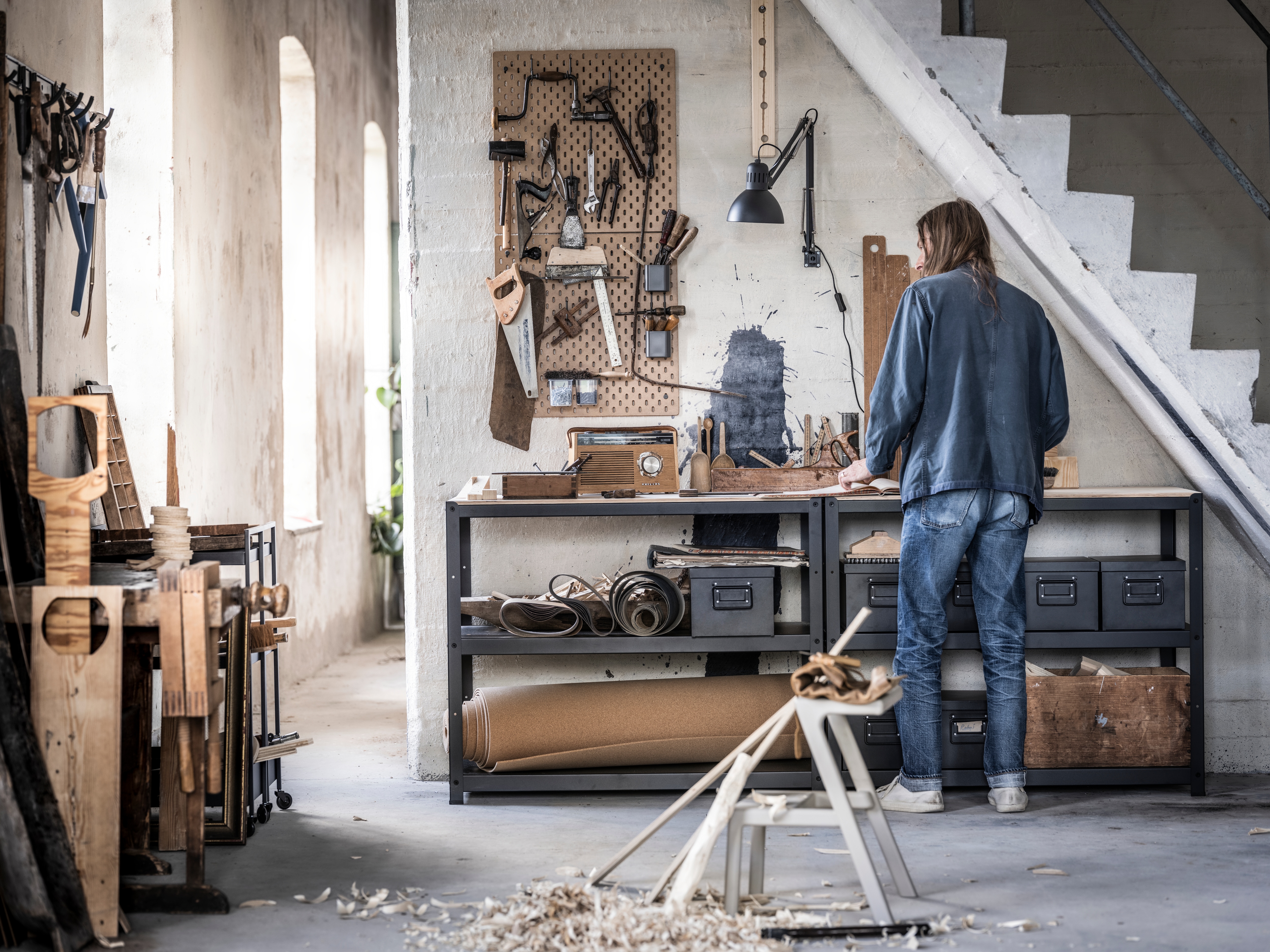 Woman standing at BROR workbenches under the stairs. SKÅDIS pegboard with tools hanging on the wall.