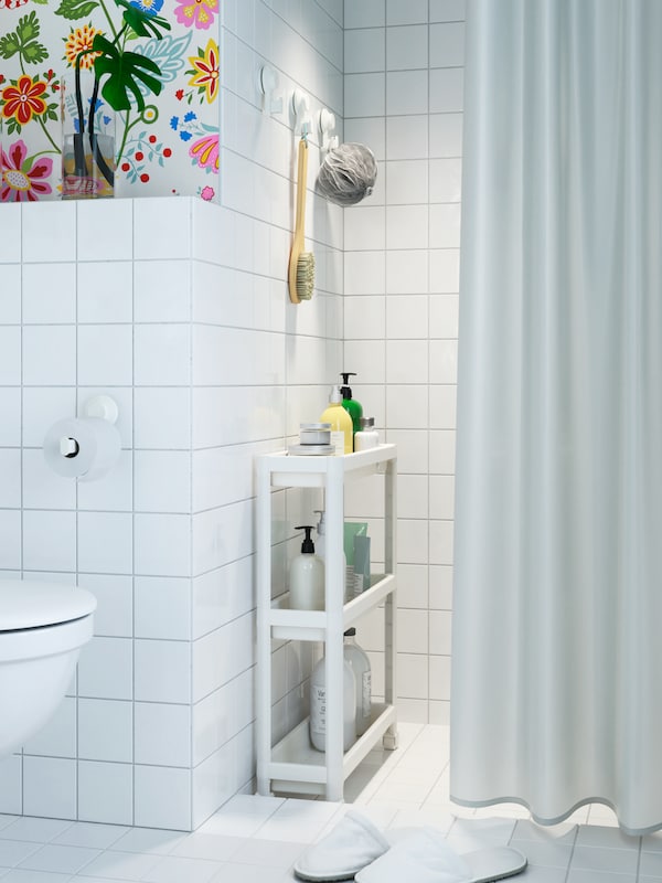 Behind a white shower curtain, a white narrow trolley with toiletries stands in the corner of the shower area.