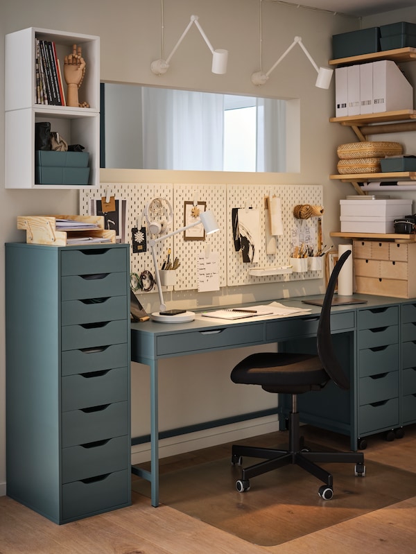 An IKEA ALEX blue desk with matching drawer units on either side, desk accessories, a black office chair and shelves on the wall.