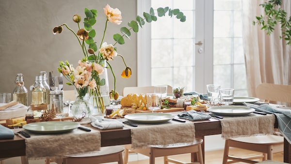 A long MÖRBYLÅNGA table festively set with lengths of AINA fabric, FÄRGKLAR plates, flowers in vases and various decorations.