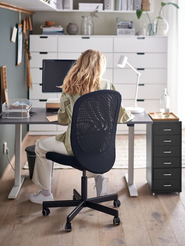 A person sitting on an IKEA FLINTAN chair in black, by a grey desk and a black drawer unit in a sunny office.