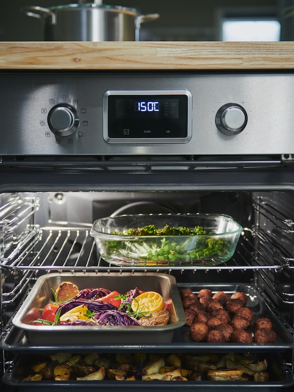 An open stainless steel SMAKSAK oven with a FÖLJSAM glass oven dish, HEMMABAK roasting tin and other trays with food inside.
