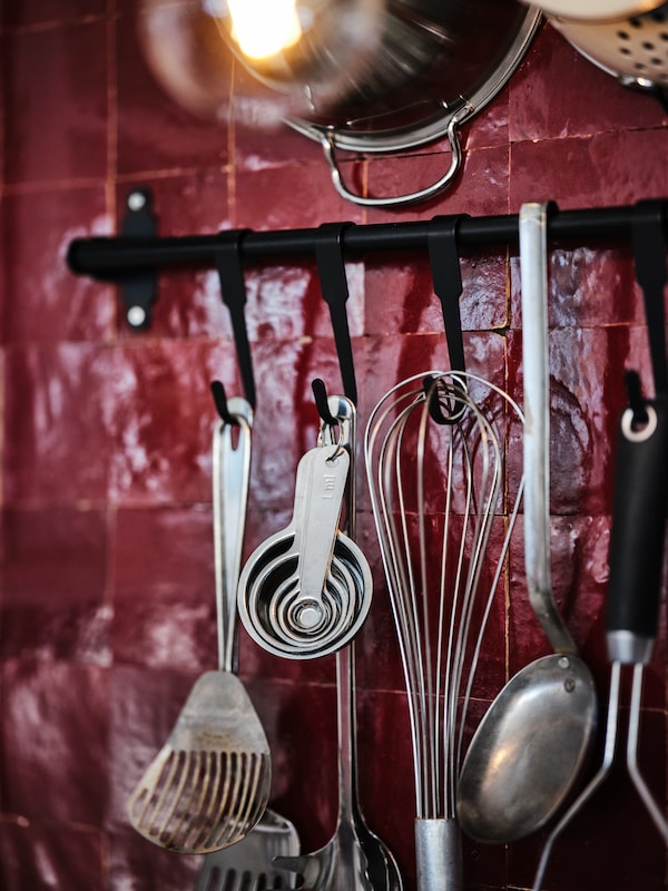 Various IKEA KONCIS cooking tools in stainless steel hung on a red tilled wall.