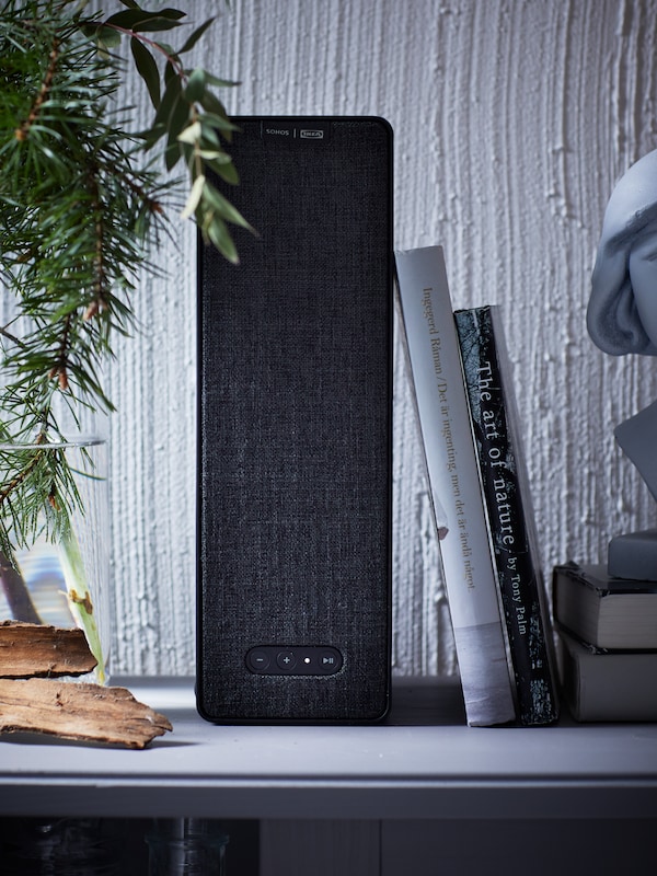 A black SYMFONISK WiFi bookshelf speaker is standing on a grey surface against a white wall. Two books are leaning on it.