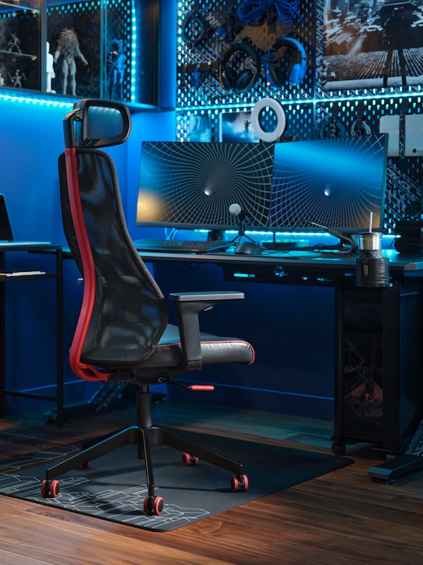 A dark gaming room, illuminated in blue, with a MATCHSPEL gaming chair on a floor protector and two screens on a gaming desk.