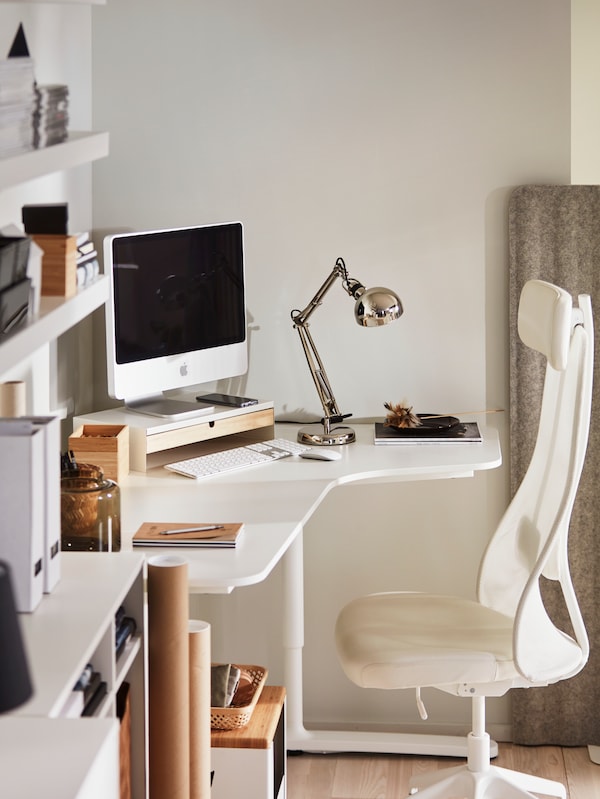 An IKEA BEKANT corner white desk with a computer, light and other desk accessories on top. A white and beige office chair.