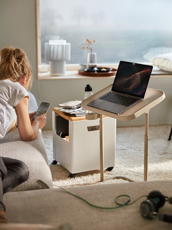 A woman is on her phone laying on the sofa. In front of her is a BJÖRKÅSEN laptop stand and TROTTEN storage unit on castors.