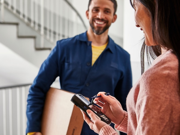 A delivery man in a blue jacket hands something to sign to a woman wearing a grey jumper. Next to her are flatpack boxes and an IKEA blue bag.