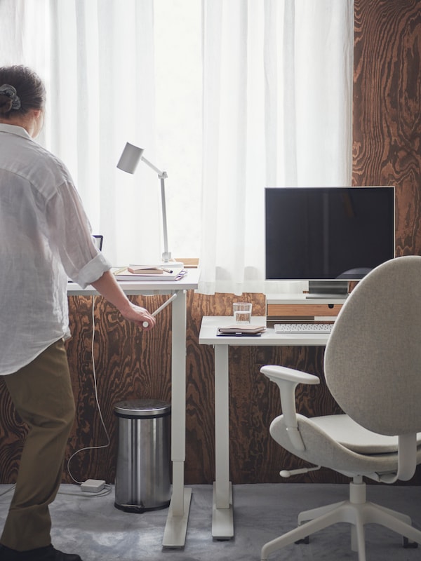Two IKEA TROTTEN sit and stand desks in white in a sunny room, a person using the handle to raise one of the desks higher up