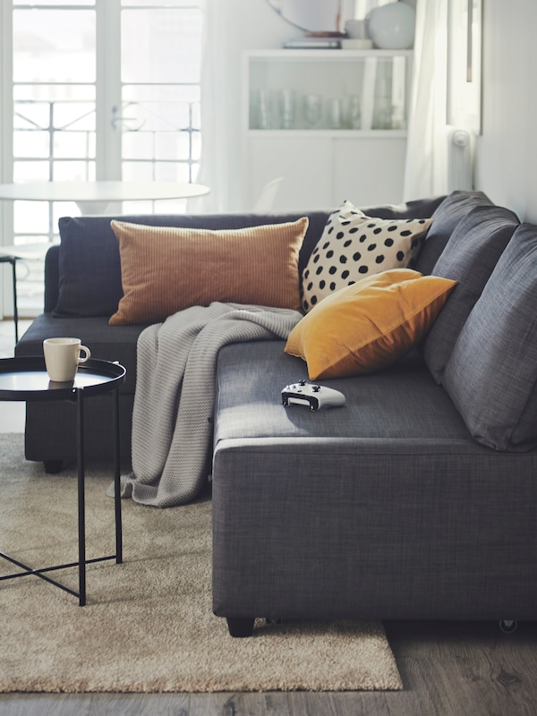 An IKEA FRIHETEN corner fabric sofa in dark grey, with cushions and a blanket thrown over, in a sunny living room.