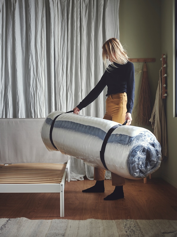 A person stands over a bed frame with a rolled up VÄSTEROY mattress. Behind them is a curtain and hooks with clothing.