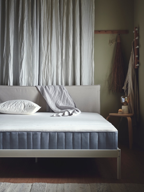 A VÄSTEROY mattress is on a bed frame with a pillow on it and blanket hanging over the headboard. A curtain hangs behind.
