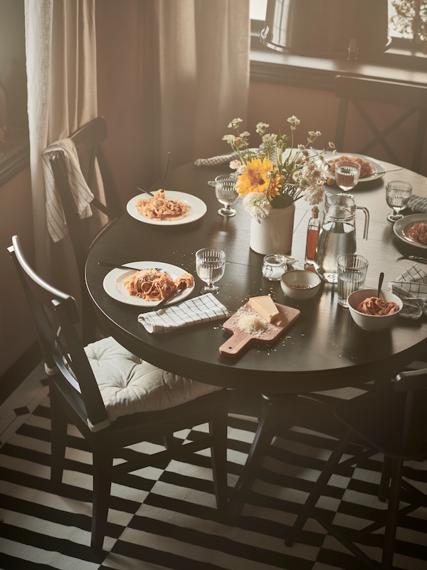 A round INGATORP dining table in black brown colour, with matching dining chairs, food, flowers and plates on the table, in front of a sunny window.