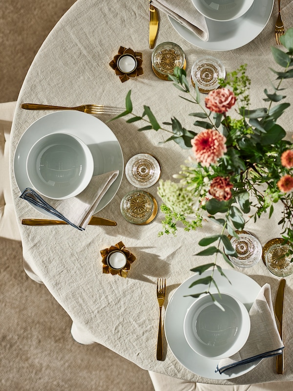 A round table with a natural OMBONAD tablecloth set with white IKEA 365+ plates and bowls, brass-colour cutlery plus flowers.