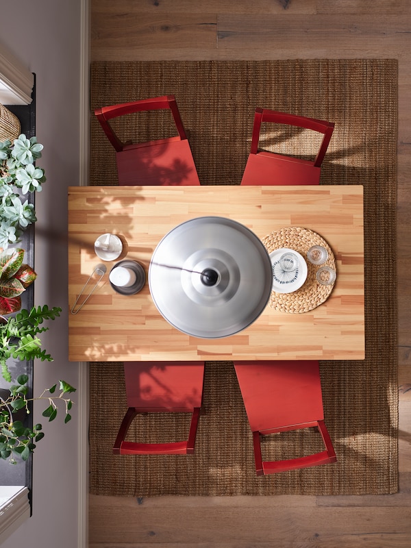 A wooden table and four red-stained PINNTORP chairs on a natural LOHALS rug under an aluminium-coloured TRETTIOEN pendant.