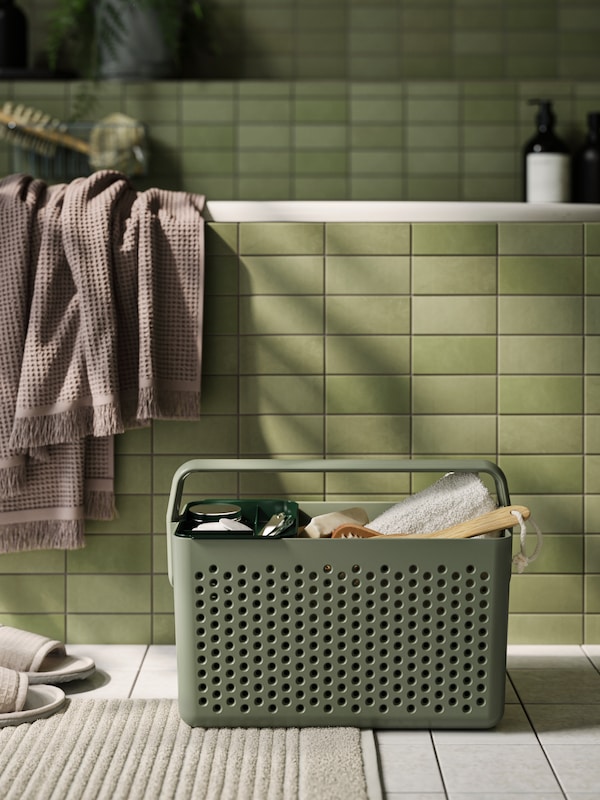 A grey-green UPPRÄMEN storage basket filled with body care items is standing on a bathroom floor in front of a green bathtub.