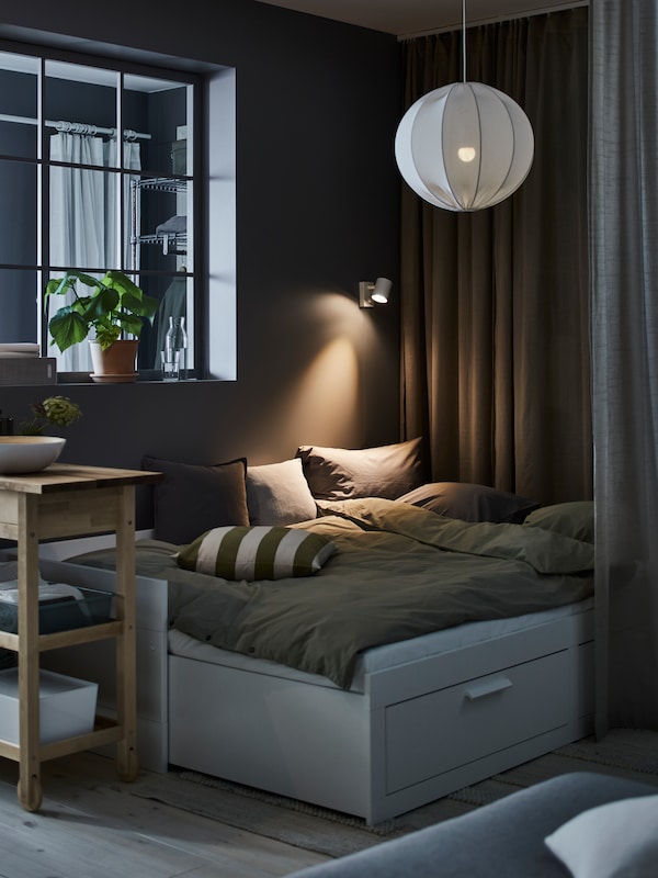 A BRIMNES day-bed with 2 drawers with bedding on it stands in a dark room under a lit REGNSKUR pendant lamp.