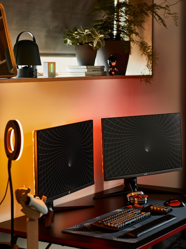 A gaming room with two screens on a HUVUDSPELARE gaming desk. The wall is illuminated in red and yellow behind the screens.