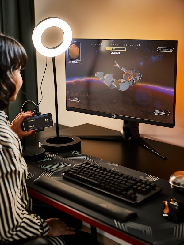 A LÅNESPELARE ring light is illuminating a person sitting behind a HUVUDSPELARE gaming desk, holding a mobile device.