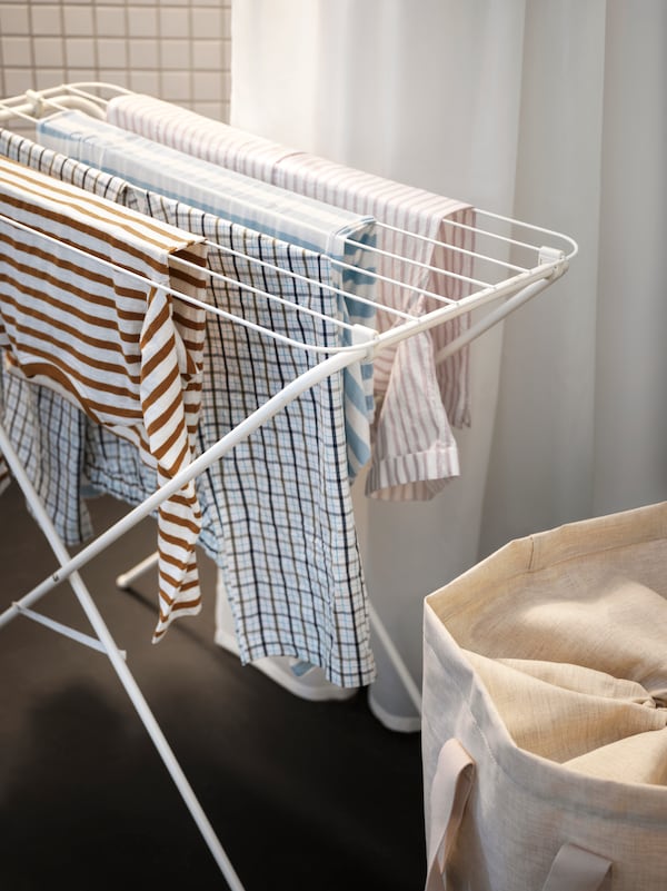 A JÄLL drying rack, in/outdoor with some patterned clothes hanging from it and a white curtain in the background.