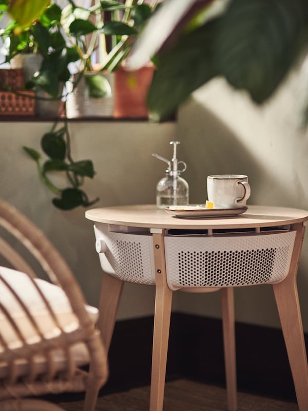 A STARKVIND table with air purifier, with a mug, a plate and a SESAMFRÖN plant mister on top of it, stands in a corner.