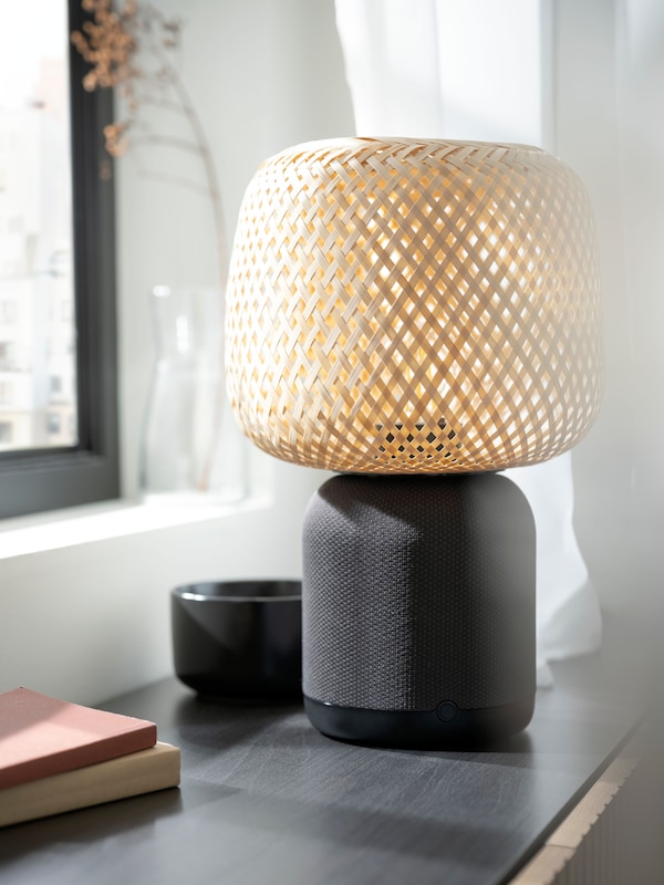 A black SYMFONISK speaker lamp base with a bamboo shade is standing on a broad black windowsill in a bright white room.