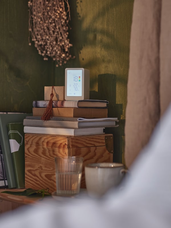 A VINDSTYRKA air quality sensor stands on top of some books on top of a wooden box beside a glass and a mug in a corner.