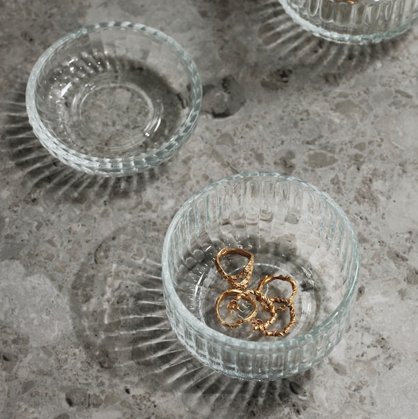 A close-up of a SILVTJÄRN glass container with a lid, placed on a grey table, with small pieces of jewellery inside.