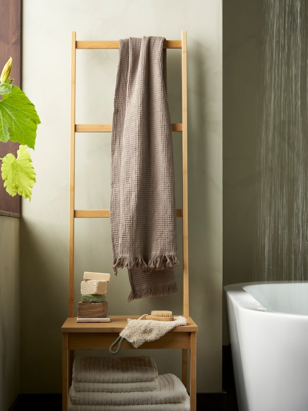VÅGSJÖN beige towels hanging from TISKEN hooks on a bathroom wall along with ÅBYÅN body puffs handing from a wall cabinet with TACKAN soap dispenser