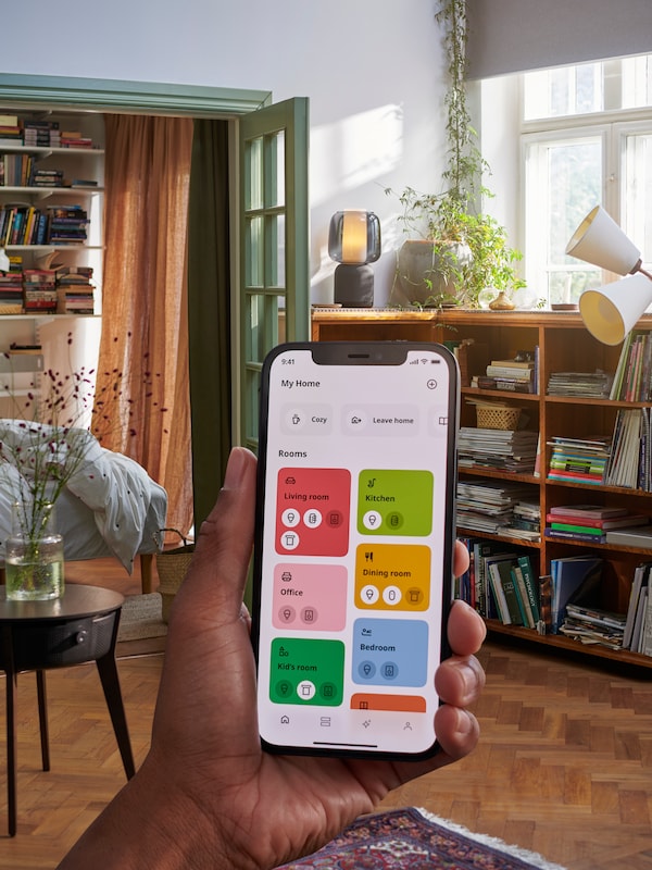 A hand holding up the IKEA Home smart app in front of a living room showing a screen with different options.
